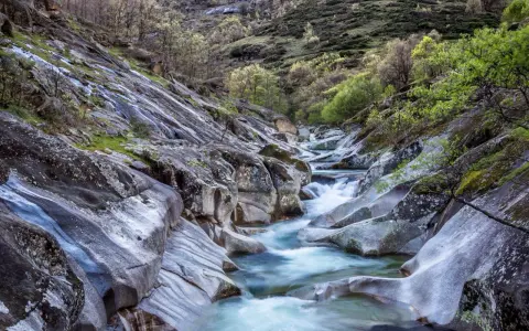 Un río de aguas cristalinas serpentea entre rocas pulidas y vegetación exuberante. Un paisaje idílico que evoca la frescura del verano en Extremadura.