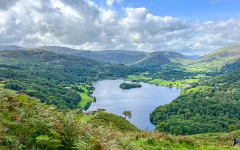 A stunning view of a lake surrounded by green hills and trees, perfect for day trips from Liverpool. A lone hiker walks down a path in the foreground.