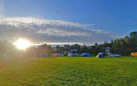 Słońce przebija się przez chmury nad polem namiotowym. Rozstawione namioty i samochody zapowiadają udany camping.