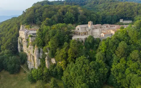 A monastery complex clings to a sheer cliff face in La Verna, Italy, surrounded by lush green forests.
