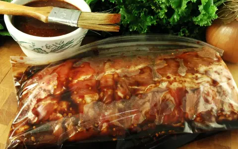 Karkówka w torebce, gotowa na grilla. W miseczce czeka aromatyczna marynata do karkówki na grilla, a obok pęczek zielonej sałaty i cebula.