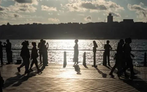 Ludzie spacerują i łowią ryby na promenadzie nad morzem. Słońce odbija się w wodzie, tworząc złoty blask.