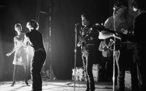 Scène de concert des membres de The Velvet Underground. Une femme danse à gauche, tandis que Lou Reed chante au micro.