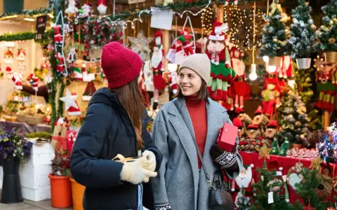 Två kvinnor njuter av julmarknader i Skåne, omgivna av julpynt och ljus. De håller presenter och ler mot varandra.