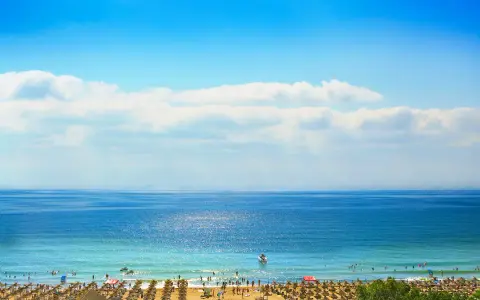 Słoneczny brzeg w pełni lata. Błękitne morze, plaża pełna ludzi i parasoli, a na horyzoncie statki. Idealna pogoda na wypoczynek.