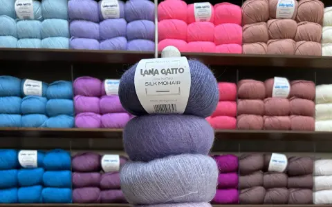 En stapel med tre härvor av lyxigt mohair silke garn i olika lila nyanser. Bakom syns hyllor fyllda med garn i många färger.