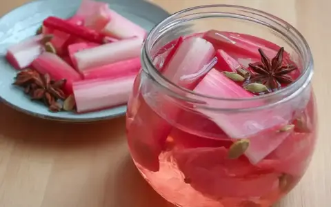 Rabarbersaft recept: färska rabarberbitar i en glasburk med kryddor som stjärnanis och kardemumma. En tallrik med rabarber och kryddor står bredvid.