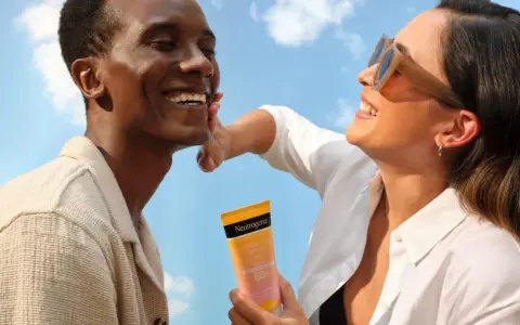 Un couple rit sous un ciel bleu, une femme appliquant une crème solaire Neutrogena. Une liste de protection contre le soleil pour une peau saine.