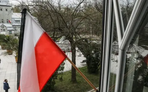 Biało-czerwona flaga na tle drzew i budynków. Czy flaga może wisieć cały rok? Pytanie o symbol dumy narodowej.