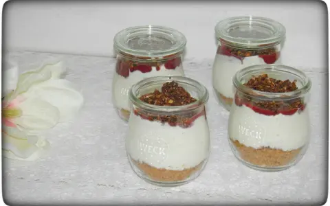 Vier leckere Desserts im Glas mit Krümelboden, cremiger Füllung, roter Fruchtsauce und knusprigem Topping.
