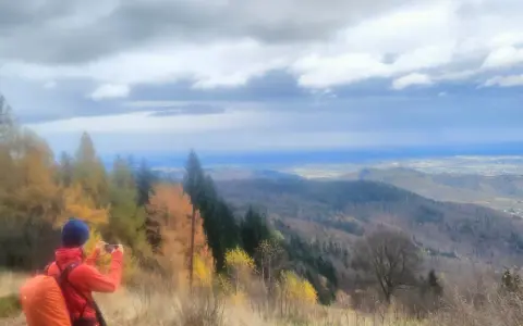 Widok na Beskid Mały jesienią. Turysta z pomarańczowym plecakiem fotografuje panoramę.