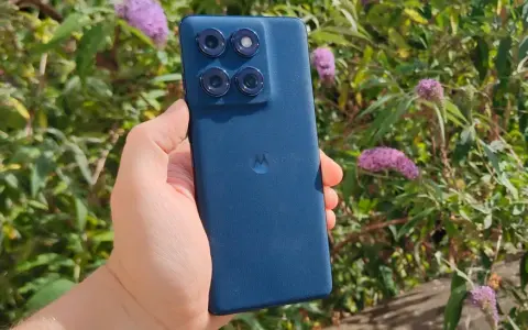 En hand håller en mörkblå motorola edge 60 fusion med fyra kameror mot en bakgrund av gröna blad och lila blommor.