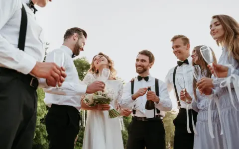 Pan młody i świadek świętują z panną młodą i druhnami. Wszyscy trzymają kieliszki, a jeden z panów otwiera butelkę szampana.