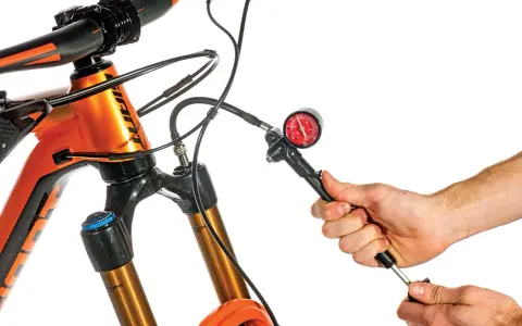Adjusting the mtb suspension setup. A hand uses a shock pump with a gauge to set the air pressure on an orange mountain bike's front fork.