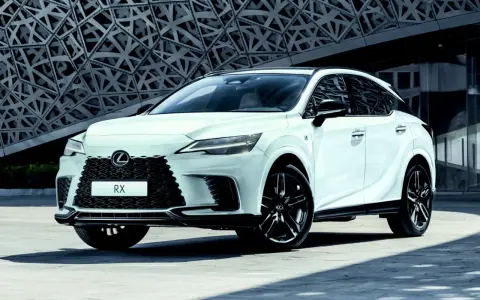 Biały Lexus RX500h z czarnymi felgami stoi przed nowoczesnym budynkiem o geometrycznej fasadzie.