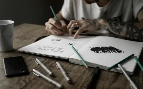 Un artiste auteur ou micro entrepreneur dessine dans son carnet, entouré de stylos, crayons et d'une tasse.