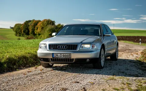 Silberner Audi A8 D2 W12 auf Schotterweg, umgeben von grünen Feldern und Bäumen im Herbstlicht.