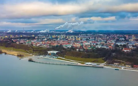 Widok na Płock z lotu ptaka, z Wisłą w tle. Widać panoramę miasta, fabryki z dymiącymi kominami i nowoczesną infrastrukturę nad rzeką, która może być częścią atrakcji dla dzieci w Płocku.