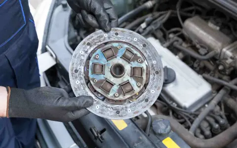 Mechanik w czarnych rękawiczkach trzyma zużyte sprzęgło samochodowe na tle silnika.