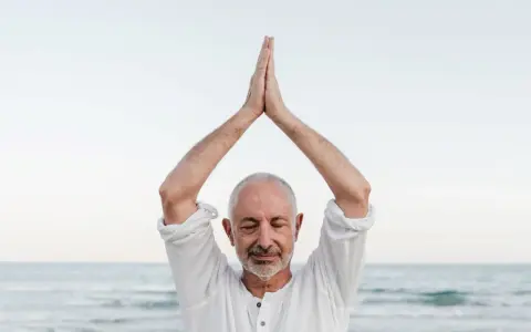 Mężczyzna z zamkniętymi oczami, ręce złożone nad głową, praktykuje mindfulness co to. Spokój nad morzem.