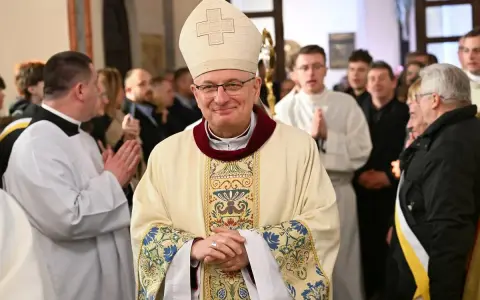 Uśmiechnięty biskup w stroju pontyfikalnym, otoczony przez wiernych i duchownych. W tle widać fragmenty kościoła, a na pierwszym planie szczegóły szat.