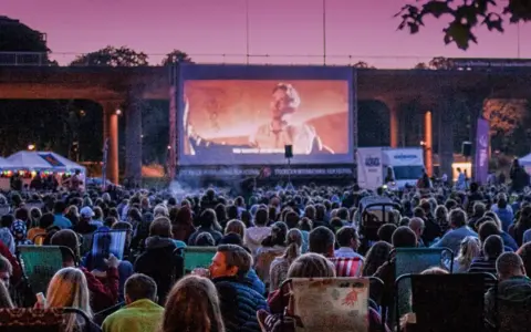 Stor publik njuter av utomhusbio i Stockholm under en rosa kvällshimmel. En stor duk visar en filmscen.