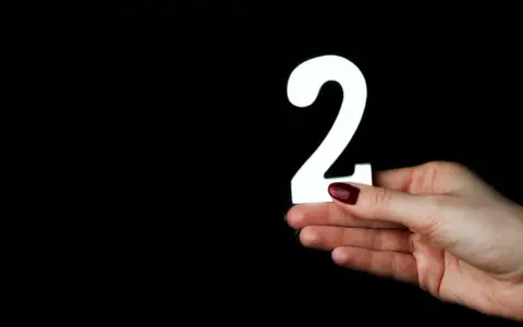 Dłoń z ciemnoczerwonym lakierem trzyma białą cyfrę 2. Co oznacza liczba 2?