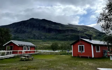 Två röda stugor i Abisko Nikkaluokta med en bergig, molntäckt bakgrund. En picknickbänk står på gräsmattan.