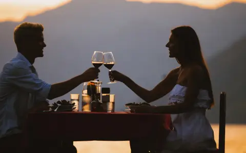 Idealne miejsce na pierwszą randkę: para wznosi toast winem przy romantycznej kolacji na tle zachodzącego słońca i gór.