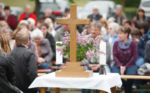 Holzkreuz mit Blumen und Kerzen vor einer Gemeinde. Ein Thema, das auch bei unterschiedlichen Konfessionen von Ehepartnern relevant ist: die Kirchensteuer.