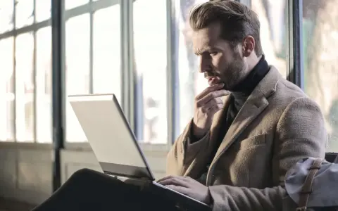Mężczyzna w płaszczu zastanawia się, jak się wymeldować z hotelu, przeglądając coś na laptopie.