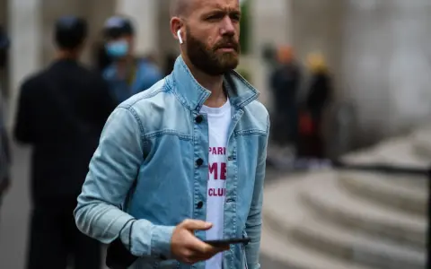 Hombre calvo sin barba, con auriculares y chaqueta vaquera, mira su móvil.