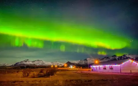Islandia pogoda listopad: zorza polarna nad ośnieżonymi górami i domkiem z lampkami.
