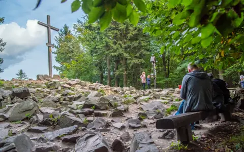 Kamienista ścieżka w **Górach Świętokrzyskich** prowadzi do drewnianego krzyża. Ludzie odpoczywają na ławkach wśród drzew.