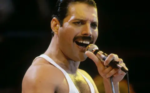 Freddie Mercury śpiewa do mikrofonu, w białej podkoszulce. Wiele osób wspomina go, kiedy zmarł Freddie Mercury.