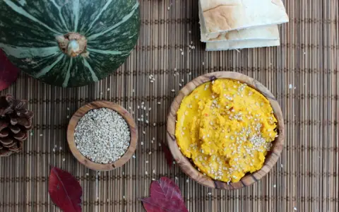 Ein leuchtend gelber Hummus, bestreut mit Sesam, wartet neben einer Schale voller Sesam und einem Kürbis. Perfekt für ein kleines Hummus-Baby-Festmahl!