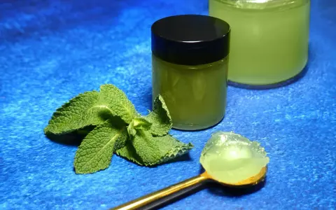 Une cuillère dorée présente une portion de gelée de menthe fraîche. À côté, un pot fermé et un autre ouvert contiennent cette même gelée verte et rafraîchissante.