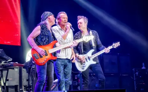 Trzech muzyków na scenie w głębokim fioletowym świetle Atlas Areny. Jeden śpiewa, dwaj grają na gitarach.