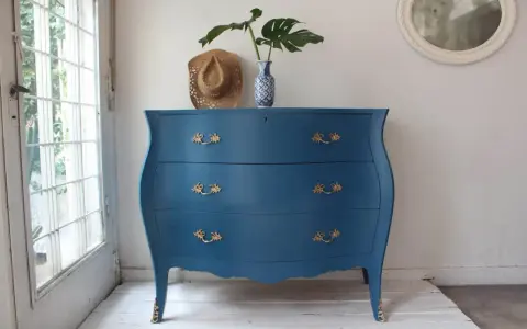 Cómoda antigua con **muebles pintados en azul** y tiradores dorados, decorada con un sombrero y un jarrón con hojas.