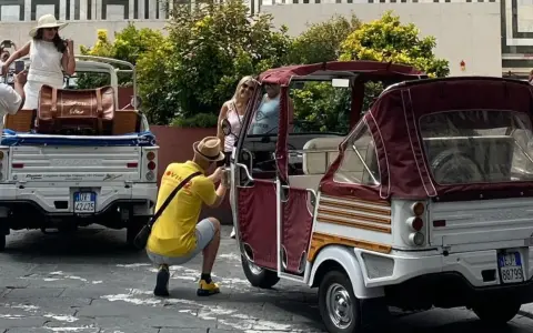 Turisti scattano foto a veicoli atipici, un Piaggio Ape e un tuk-tuk, in una strada soleggiata.