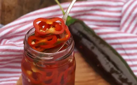 En burk med picklad chili, med en sked som lyfter upp en skiva. En mörk chili ligger bredvid.