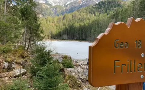 Holzschild "Geo 18 Fritter" vor gefrorenem See und Bergkulisse. Perfekt für einen Sommerurlaub in Bayern.
