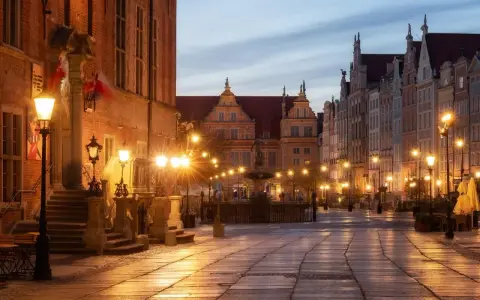 Wieczorny rynek Gdańsk oświetlony latarniami, z fontanną i zabytkowymi kamienicami.