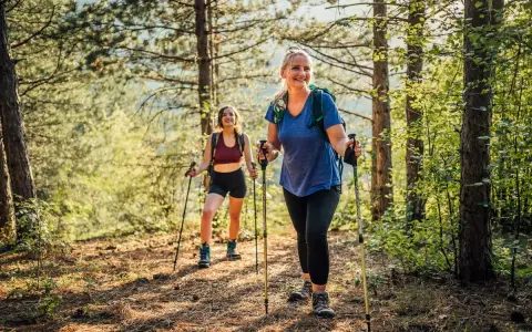 Deux femmes souriantes randonnent en forêt, utilisant des bâtons. Elles semblent apprécier la nature, un argument pour ou contre les bâtons de randonnée.
