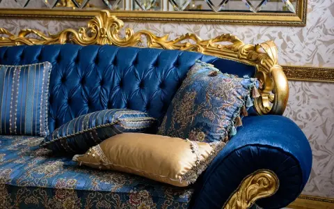 Canapé bleu profond, capitonné, aux ornements dorés, style baroque. Coussins aux motifs floraux et rayés, franges et pompons ajoutent une touche luxueuse.