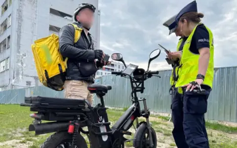 Dostawca z żółtą torbą na plecach rozmawia z policjantkami obok swojego roweru elektrycznego bez pedałowania.