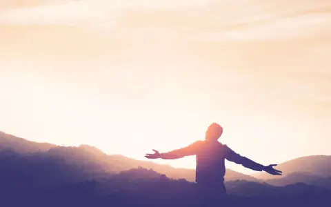 Silhouette einer Person mit ausgebreiteten Armen vor einer Bergkulisse bei Sonnenuntergang. Ein Symbol für Kraft und Hoffnung nach einer Krankheit.