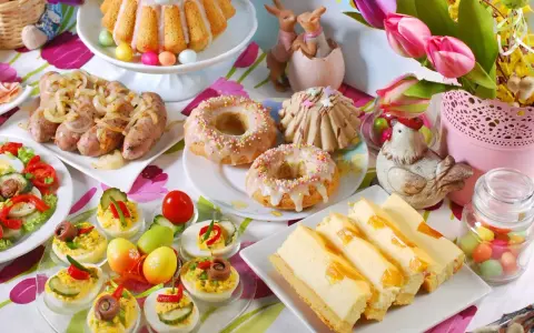 Kolorowy stół z pysznym śniadaniem wielkanocnym: babka, kiełbaski, sałatka, babeczki, sernik i jajka faszerowane.