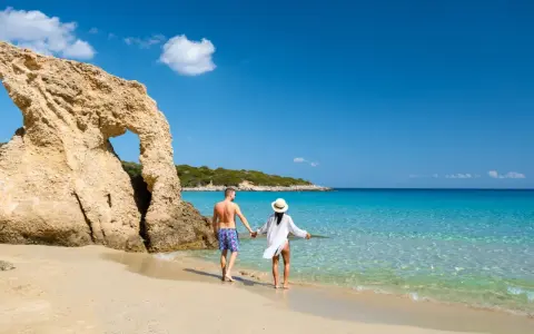 Para, trzymając się za ręce, spaceruje po piaszczystej plaży z turkusową wodą. Czy Cypr jest bezpieczny? Tak, to idealne miejsce na wakacje.