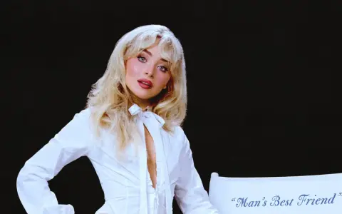 Sabrina Carpenter w białej koszuli, z rozpuszczonymi blond włosami, wygląda jak "man's best friend" na tle czerni.